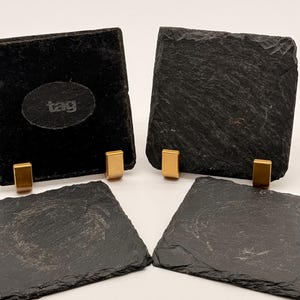May include: Four square black slate coasters with rough, textured edges. Two coasters are standing upright in gold-toned metal holders, while two are lying flat. One upright coaster features the word tag embossed in an oval center area.
