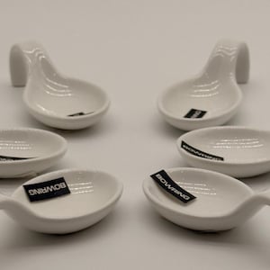Pode incluir: Um conjunto de quatro apoios de colher em cerâmica branca, cada um com um cabo curvo e uma taça oval rasa. Cada peça tem uma pequena etiqueta retangular preta colada no interior com o texto branco BOWRING impresso.