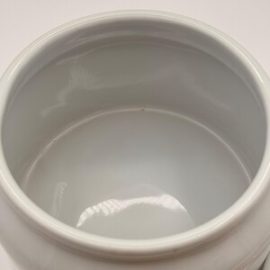 May include: A white ceramic jar with a smooth, glossy finish and a wide, circular opening. The interior of the container is deep and hollow, featuring a clean, uniform white surface throughout the basin and the rounded rim of the vessel.
