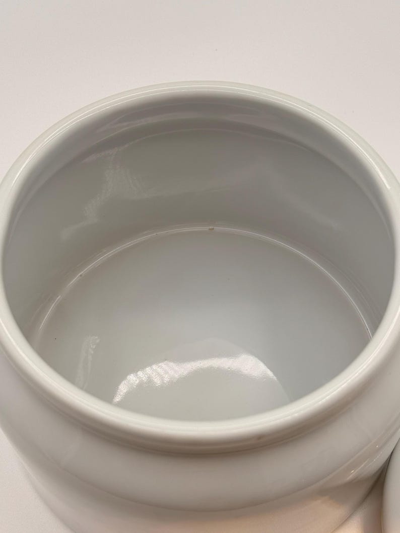May include: A white ceramic jar with a smooth, glossy finish and a wide, circular opening. The interior of the container is deep and hollow, featuring a clean, uniform white surface throughout the basin and the rounded rim of the vessel.