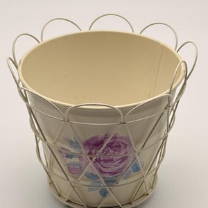 May include: A cream-coloured metal planter bucket featuring a painted purple rose design on the front. The bucket is encased in a decorative cream wire cage frame that extends above the rim in a series of rounded loops and forms a diamond pattern.