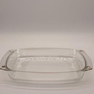 Peut inclure: Un plat de cuisson rectangulaire en verre transparent avec des coins arrondis et des poignées intégrées sur deux côtés. Le plat présente une surface lisse et transparente ainsi qu'un bassin peu profond, conçu pour une utilisation au four.