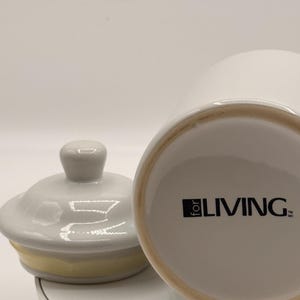 Puede incluir: Un tarro de cer&aacute;mica blanco con tapa y una banda amarilla. El tarro tiene el texto "for LIVING" impreso en negro. La tapa est&aacute; a un lado, revelando la abertura del tarro. El tarro est&aacute; sobre una superficie blanca.