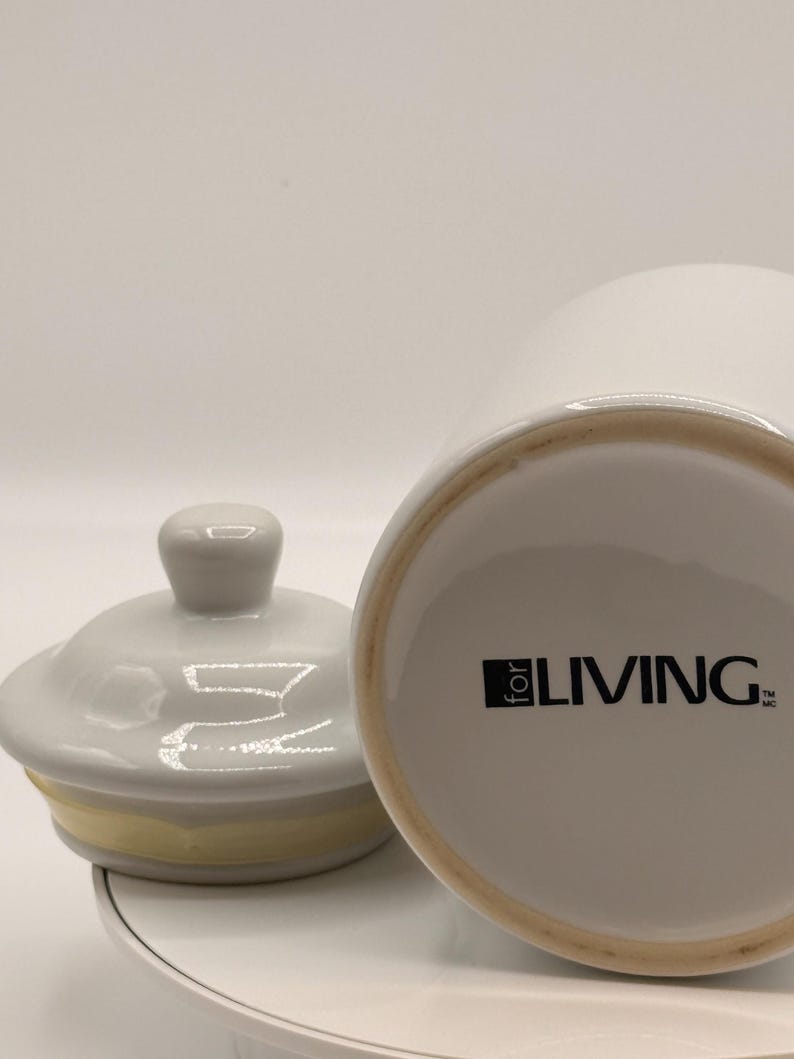 Puede incluir: Un tarro de cer&aacute;mica blanco con tapa y una banda amarilla. El tarro tiene el texto "for LIVING" impreso en negro. La tapa est&aacute; a un lado, revelando la abertura del tarro. El tarro est&aacute; sobre una superficie blanca.