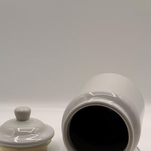 Puede incluir: Un tarro de cer&aacute;mica gris de dos tonos con tapa. El tarro es cil&iacute;ndrico con una abertura ancha, y la tapa es redonda con un peque&ntilde;o pomo. El interior del tarro es negro.