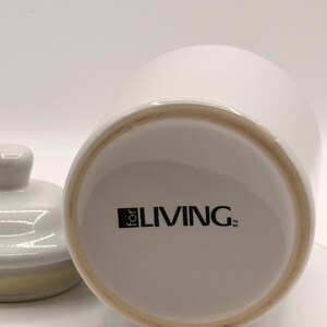 Puede incluir: Un tarro de cer&aacute;mica blanco con tapa, sobre un fondo blanco. La base del tarro presenta el texto "for LIVING" en negro. La tapa est&aacute; a un lado, revelando el interior del tarro.