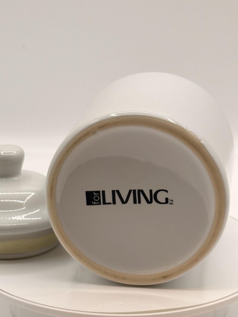 Puede incluir: Un tarro de cer&aacute;mica blanco con tapa, sobre un fondo blanco. La base del tarro presenta el texto "for LIVING" en negro. La tapa est&aacute; a un lado, revelando el interior del tarro.