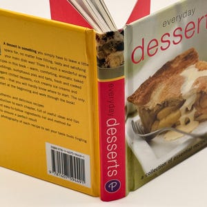 May include: A cookbook titled Everyday Desserts with a yellow back cover featuring text and a barcode, a red spine with white text, and a front cover showing a slice of apple pie with cream and a fork. The book is standing open with pages visible.