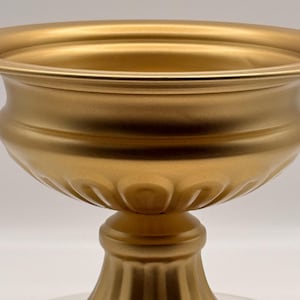 May include: A gold-coloured, pedestal urn with a wide, shallow bowl and a fluted base. The urn has a classic, elegant design, suitable for floral arrangements or decorative display. The metallic finish adds a touch of luxury.