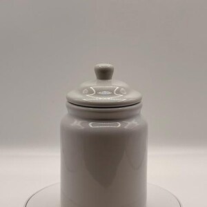 Puede incluir: Un tarro de cer&aacute;mica gris con tapa. El tarro es cil&iacute;ndrico con una parte superior redondeada y un peque&ntilde;o pomo en la tapa. El tarro est&aacute; sobre una superficie blanca.