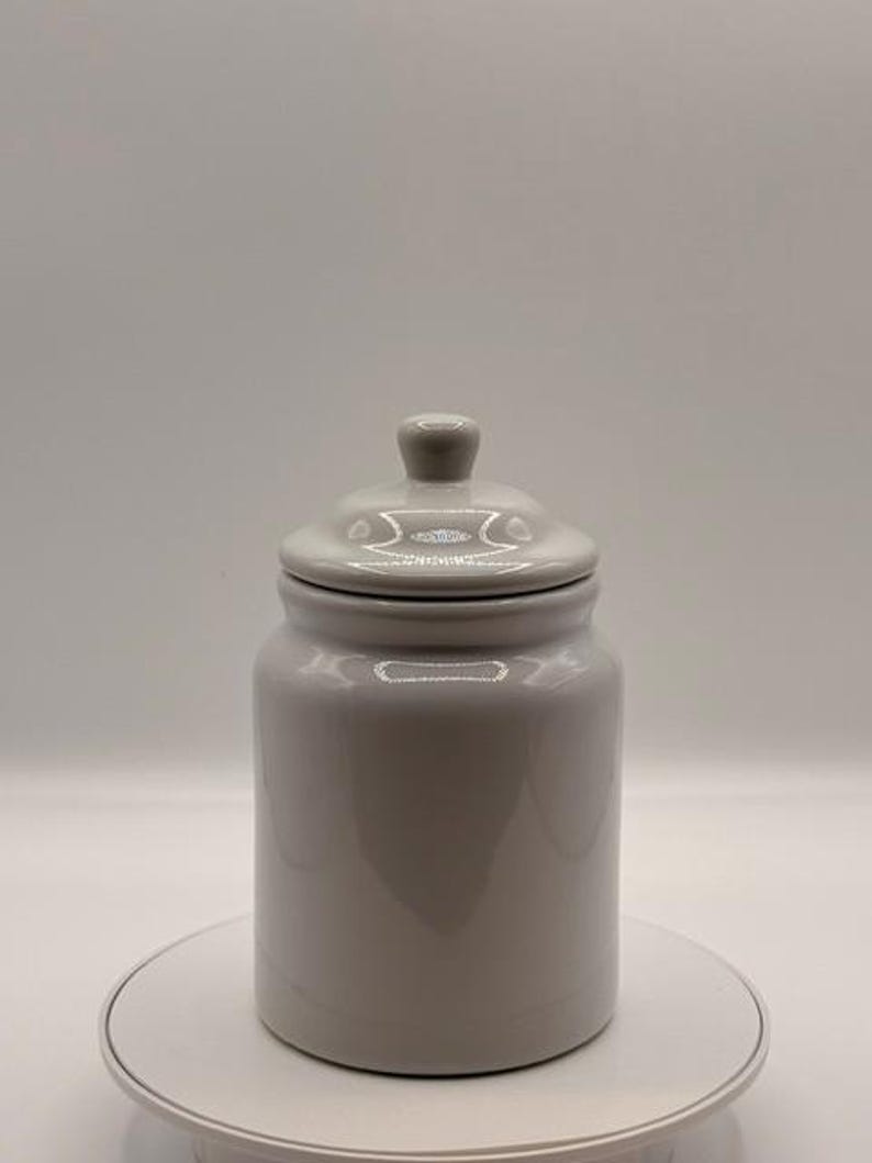 Puede incluir: Un tarro de cer&aacute;mica gris con tapa. El tarro es cil&iacute;ndrico con una parte superior redondeada y un peque&ntilde;o pomo en la tapa. El tarro est&aacute; sobre una superficie blanca.