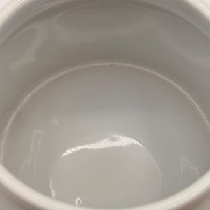 May include: A white ceramic bowl with a smooth, glossy interior finish and a rounded rim. The bowl features a deep, open basin with a curved base, reflecting light on its polished surface. The design is simple, functional, and uniform in color.