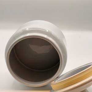 May include: A white ceramic canister with a smooth, glossy finish and a wide, open top. Beside the canister sits a matching white lid featuring a small, rounded handle and a yellow silicone gasket seal around the inner rim for a secure closure.
