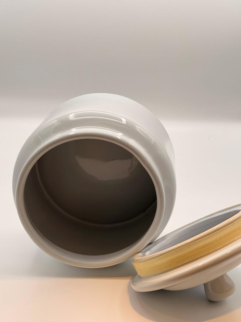 May include: A white ceramic canister with a smooth, glossy finish and a wide, open top. Beside the canister sits a matching white lid featuring a small, rounded handle and a yellow silicone gasket seal around the inner rim for a secure closure.