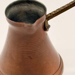 May include: A copper coffee pot with a wide base, a narrow neck, and a flared rim featuring a small pouring spout. The pot has a long, textured metal handle attached to the side with a rivet. The interior of the pot is metallic silver.