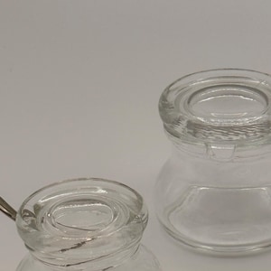 May include: Two clear glass jars with rounded bodies and wide openings. One jar features a small metal spoon resting inside, while the other jar is empty. Both containers have thick, smooth glass rims and a transparent, colorless appearance.