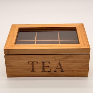 May include: A rectangular bamboo tea storage box featuring a hinged lid with a clear glass window. The front panel of the box is engraved with the word TEA in a dark, serif font. The interior is divided into six compartments for organisation.