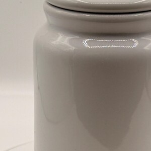 Puede incluir: Un tarro de cer&aacute;mica blanco con tapa. El tarro tiene forma cil&iacute;ndrica con un acabado liso y brillante. La tapa tiene un patr&oacute;n decorativo en el borde. El tarro probablemente se utiliza para el almacenamiento.