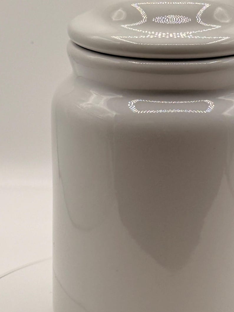 Puede incluir: Un tarro de cer&aacute;mica blanco con tapa. El tarro tiene forma cil&iacute;ndrica con un acabado liso y brillante. La tapa tiene un patr&oacute;n decorativo en el borde. El tarro probablemente se utiliza para el almacenamiento.