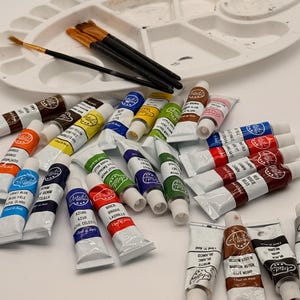 Może przedstawiać: Biała plastikowa paleta malarska z kilkoma pędzlami o czarnych trzonkach, otoczona kolekcją małych tubek z farbą o pojemności 12 ml w różnych kolorach, w tym czerwonym, niebieskim, zielonym, żółtym, brązowym, białym i czarnym.