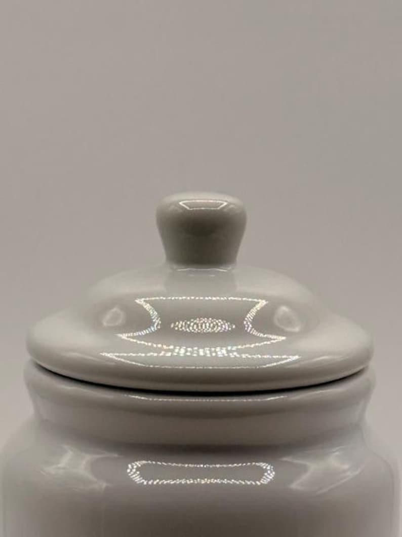 Puede incluir: Un tarro de cer&aacute;mica gris claro con tapa a juego. La tapa tiene un peque&ntilde;o pomo redondeado para facilitar su manejo. El tarro y la tapa tienen un acabado brillante, que refleja la luz. El tarro probablemente se utiliza para el almacenamiento.