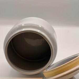 May include: A white ceramic canister with a cylindrical body and an open top. Beside the canister lies a matching white ceramic lid, which features a small knob handle and a yellow silicone gasket seal around the inner rim for a secure closure.