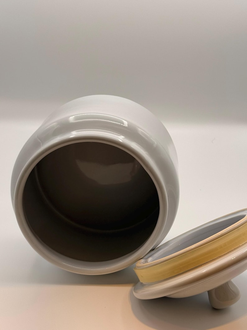 May include: A white ceramic canister with a cylindrical body and an open top. Beside the canister lies a matching white ceramic lid, which features a small knob handle and a yellow silicone gasket seal around the inner rim for a secure closure.