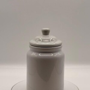 Puede incluir: Un tarro de cer&aacute;mica gris claro con tapa a juego y un peque&ntilde;o pomo en la parte superior. El tarro es cil&iacute;ndrico con una base redondeada y un acabado liso y brillante. El tarro est&aacute; sobre una superficie blanca.