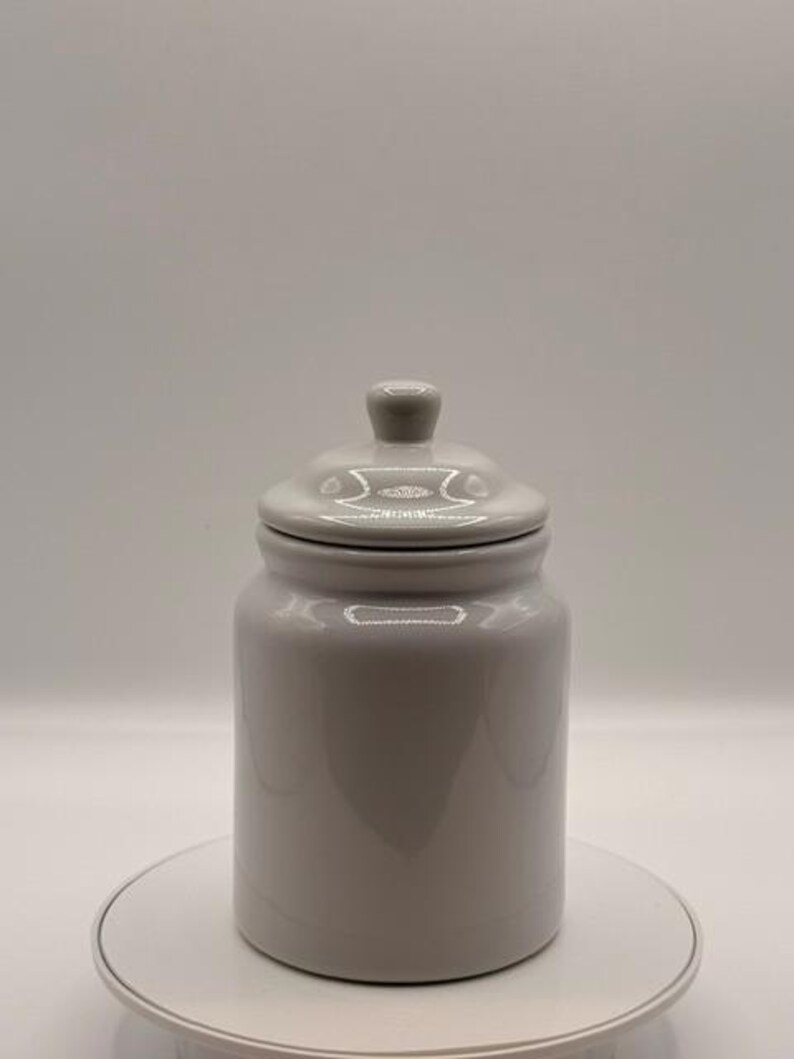 Puede incluir: Un tarro de cer&aacute;mica gris claro con tapa a juego y un peque&ntilde;o pomo en la parte superior. El tarro es cil&iacute;ndrico con una base redondeada y un acabado liso y brillante. El tarro est&aacute; sobre una superficie blanca.