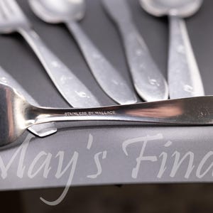 May include: A set of silver-toned stainless steel flatware including forks and spoons. The handle of the foreground fork is clearly engraved with the text STAINLESS BY WALLACE. The utensils feature a smooth, polished finish and a simple design.