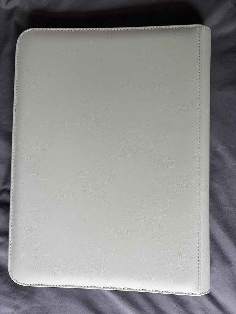 K&ouml;nnte beinhalten: Eine rechteckige, cremewei&szlig;e Dokumentenmappe mit einer glatten, lederartigen Textur. Die Au&szlig;enfl&auml;che ist einfarbig und schlicht gehalten, mit sichtbaren N&auml;hten entlang der Kanten und einem verst&auml;rkten Buchr&uuml;cken auf der rechten Seite des Einbands.