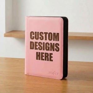 May include: A light pink rectangular binder with a dark brown spine and edges. The front cover features the text CUSTOM DESIGNS HERE in large, dark brown, block-style letters. The brand name ZOFLY is embossed in small letters at the bottom.