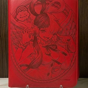 May include: A red zipped binder featuring an embossed anime-style character with long hair, headphones, and wings. The design includes a small smiling cloud and star icon within an oval frame. The Ultra PRO logo is visible in the bottom right corner.