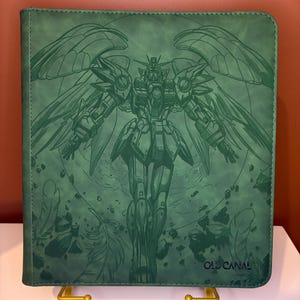 May include: A rectangular green binder featuring a dark green line art illustration of a winged robot character. The design includes floating feathers and debris surrounding the figure. The text OLD CANAL is printed in the bottom right corner.