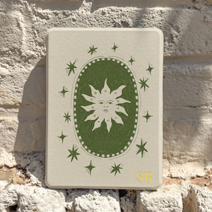 Puede incluir: Una baldosa decorativa rectangular, blanquecina, con un diseño de sol verde dentro de un marco ovalado, rodeado de estrellas. Las iniciales "SB" están en la esquina inferior derecha. La baldosa está colocada contra una pared de ladrillos blancos texturizados.