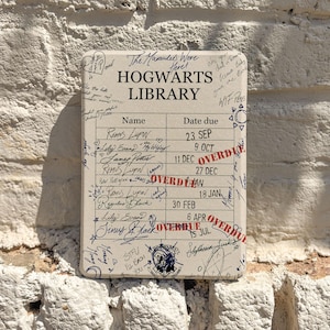 May include: A rectangular off-white sign featuring the text "The Marauders Were Here! HOGWARTS LIBRARY" above a library checkout card grid. The card includes handwritten names, dates, and red "OVERDUE" stamps, along with various doodles and signatures.