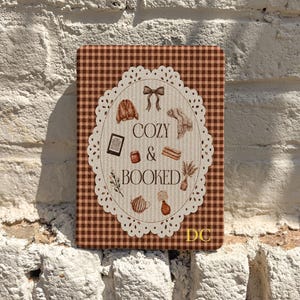 Pode incluir: Uma placa retangular com padrão xadrez castanho e branco com uma moldura oval de renda branca no centro. Dentro há ilustrações de um laço, camisola, cachecol, leitor eletrónico, caneca, livro, planta, bule e candeeiro com o texto COZY & BOOKED.