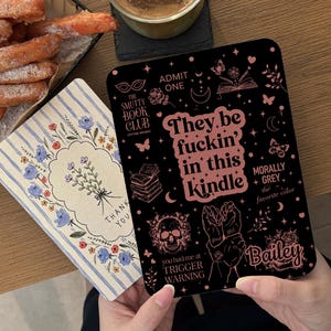 Könnte beinhalten: Eine schwarze Kindle-Hülle mit rosa Text, der "They be fuckin' in this Kindle" lautet. Die Hülle hat einen Totenkopf, Schmetterlinge und andere Grafiken. Eine florale "Thank You"-Karte und eine Tasse Kaffee sind ebenfalls sichtbar.
