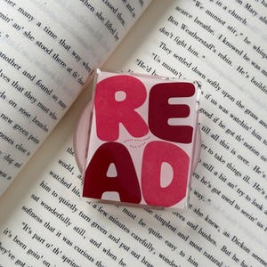 May include: A square, clear acrylic phone grip featuring the word READ in bold, blocky letters. The letters are coloured in varying shades of pink and dark red. Small text below the word reads BOOKS OPEN MINDS READ OFTEN. The grip is placed on a book.
