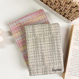 May include: Two rectangular passport holders with a woven fabric texture. One holder features a multi-colored pattern of pink, yellow, and blue threads, while the other is grey and white with the name Sophia printed in black script on the bottom right.