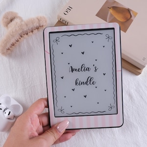 May include: An e-reader device featuring a light pink and white vertical striped frame. The screen displays the text 'Amelia's kindle' in a cursive font, surrounded by a decorative border with small heart and bow illustrations on a light gray background.