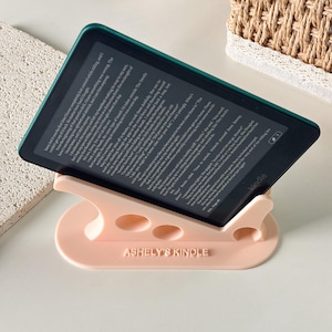May include: A black e-reader device displaying text on its screen, resting on a light pink plastic stand. The stand features three circular cutouts and the embossed text ASHELY'S KINDLE on the front base, holding the device at an angled position.