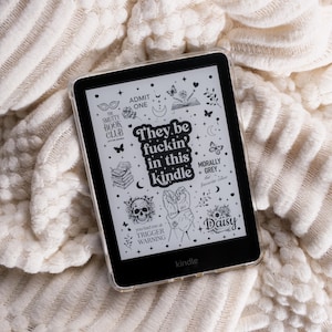 Op de afbeelding: Een zwarte Kindle e-reader met een doorzichtige beschermhoes en een witte sticker met zwarte illustraties en tekst: They be fuckin' in this kindle, The Smutty Book Club, Morally Grey, Daisy en you had me at TRIGGER WARNING.