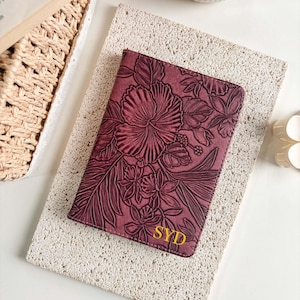 Könnte beinhalten: Ein rechteckiger, burgunderroter Reisepasshalter mit einem geprägten Blumenmuster aus Hibiskusblüten und tropischen Blättern. In der unteren rechten Ecke befinden sich die Buchstaben SYD in gelben, serifenlosen Großbuchstaben.