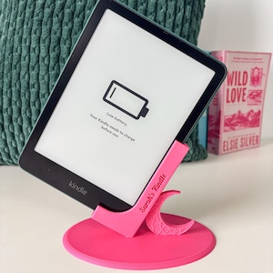 May include: A black Kindle e-reader resting on a bright pink plastic stand. The stand features a circular base, a crescent moon cutout, and the text Sarah's Kindle printed on the side. The screen displays a low battery icon and text.