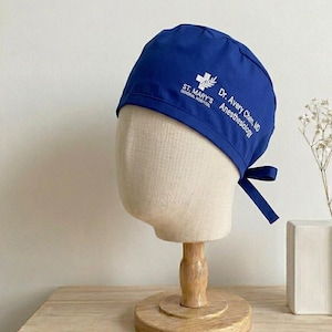 Könnte beinhalten: Eine blaue OP-Haube mit weißem Text und einem Kreuzlogo. Die Haube ist hinten gebunden und trägt den Text "St. Mary's General Hospital" und "Dr. Avery Chen, MD Anesthesiology". Die Haube wird auf einem Schaufensterpuppenkopf präsentiert.