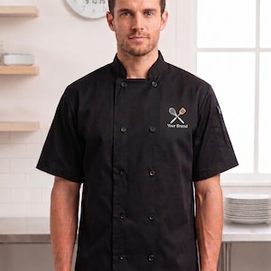 May include: A black short-sleeved chef jacket featuring a mandarin collar, a double-breasted front with ten black buttons, and a small embroidered logo on the left chest depicting a crossed whisk and spatula with the text Your Brand below it.