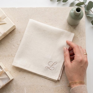 Può includere: Un tovagliolo di lino color bianco sporco di forma quadrata, caratterizzato da una delicata lettera L ricamata in corsivo con un filo color tortora chiaro. Il tessuto presenta una trama naturale visibile ed è piegato in forma quadrata.