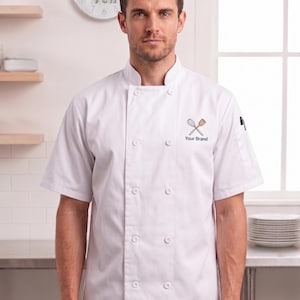 May include: A white short-sleeved chef coat featuring a mandarin collar, a double-breasted front with white buttons, and an embroidered logo on the chest depicting a crossed whisk and spatula above the text Your Brand in black lettering.