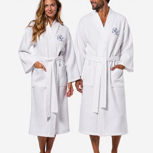 May include: Two white waffle-weave bathrobes featuring a shawl collar, a tie belt at the waist, and two front patch pockets. Each robe is embroidered on the left chest with the navy blue monogram letters RL in a cursive script font.